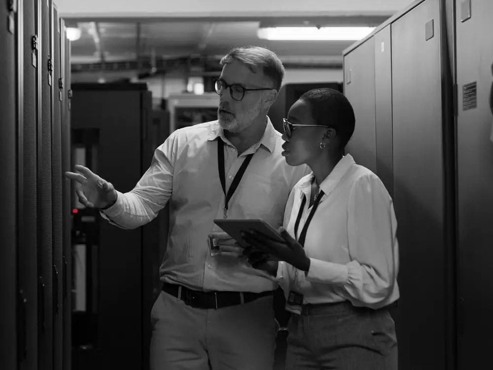 Two auditors reviewing server racks in a data center, one pointing at a rack while the other checks a tablet