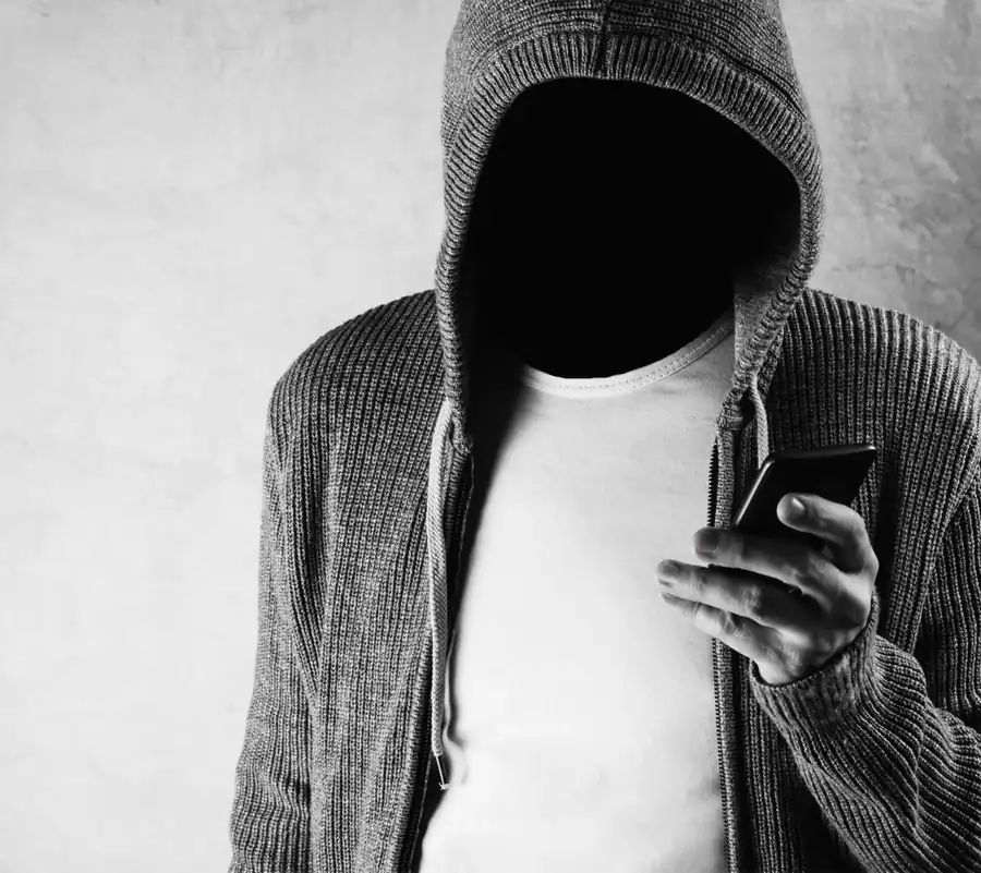 A hooded figure with a hidden face holding a smartphone, the anonymous caller or sender behind a social engineering attack