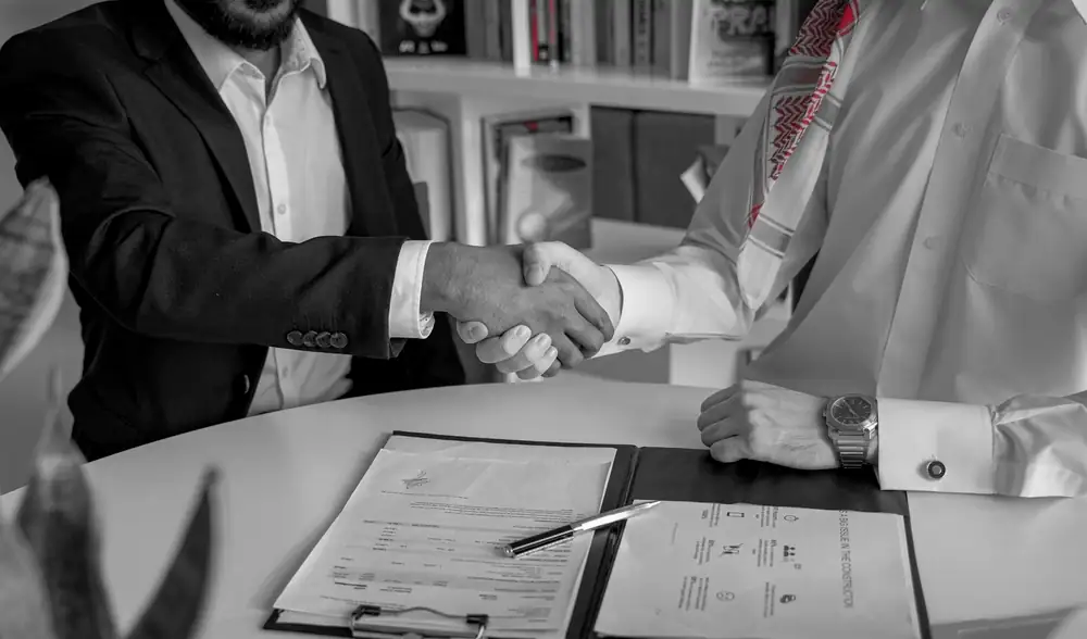 Two business leaders shaking hands over signed deal documents on a conference table